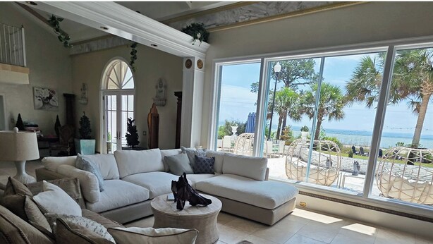 A living room with a large sectional sofa, a round coffee table, and a view of the beach through large windows. - Bay-Front Dream Mansion and Guest House -20 minutes to Destin beaches on 4 acres (Freeport)