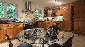 A modern kitchen with wooden cabinets, a stainless steel range hood, a glass dining table, and a view of greenery outside. - Merewood Lodge (Windermere)