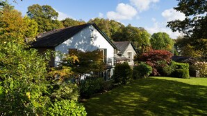 A white house with a stone roof, surrounded by lush greenery and trees. - Merewood Lodge (Windermere)