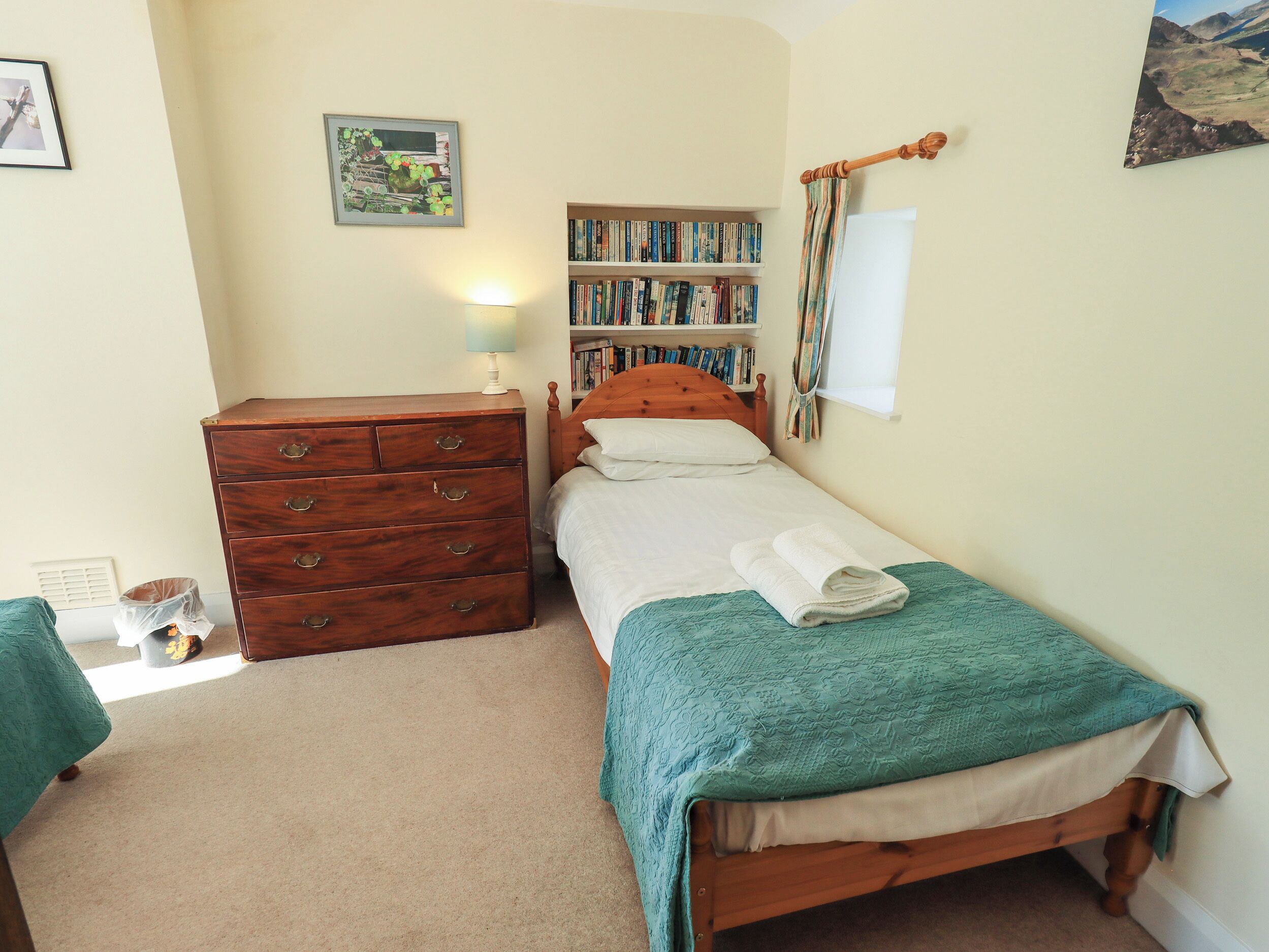 Une chambre avec deux lits simples, une commode en bois, une bibliothèque et une fenêtre avec des rideaux.