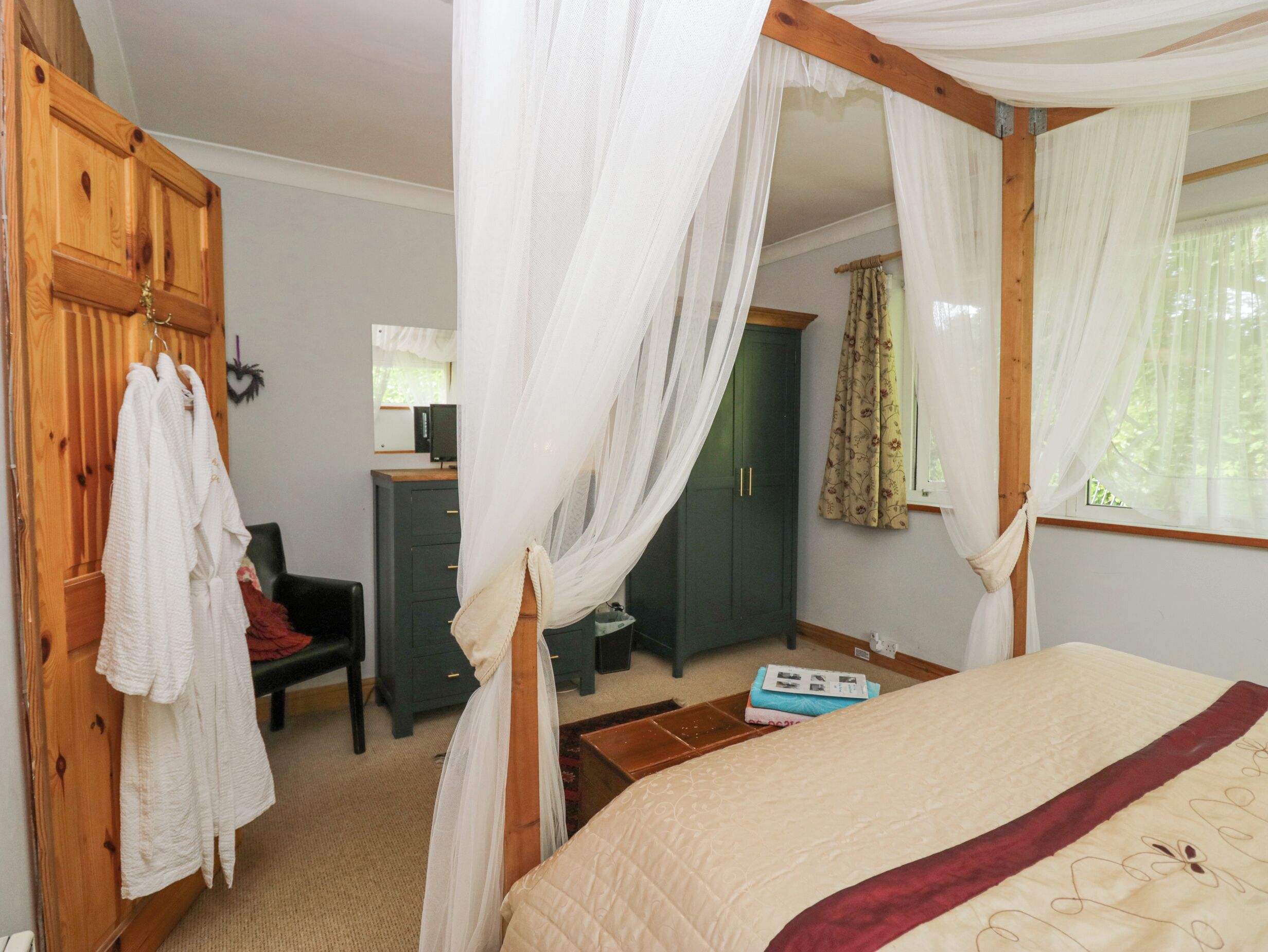 A bedroom with a four-poster bed, a wooden wardrobe, a desk with a chair, and a window with curtains.