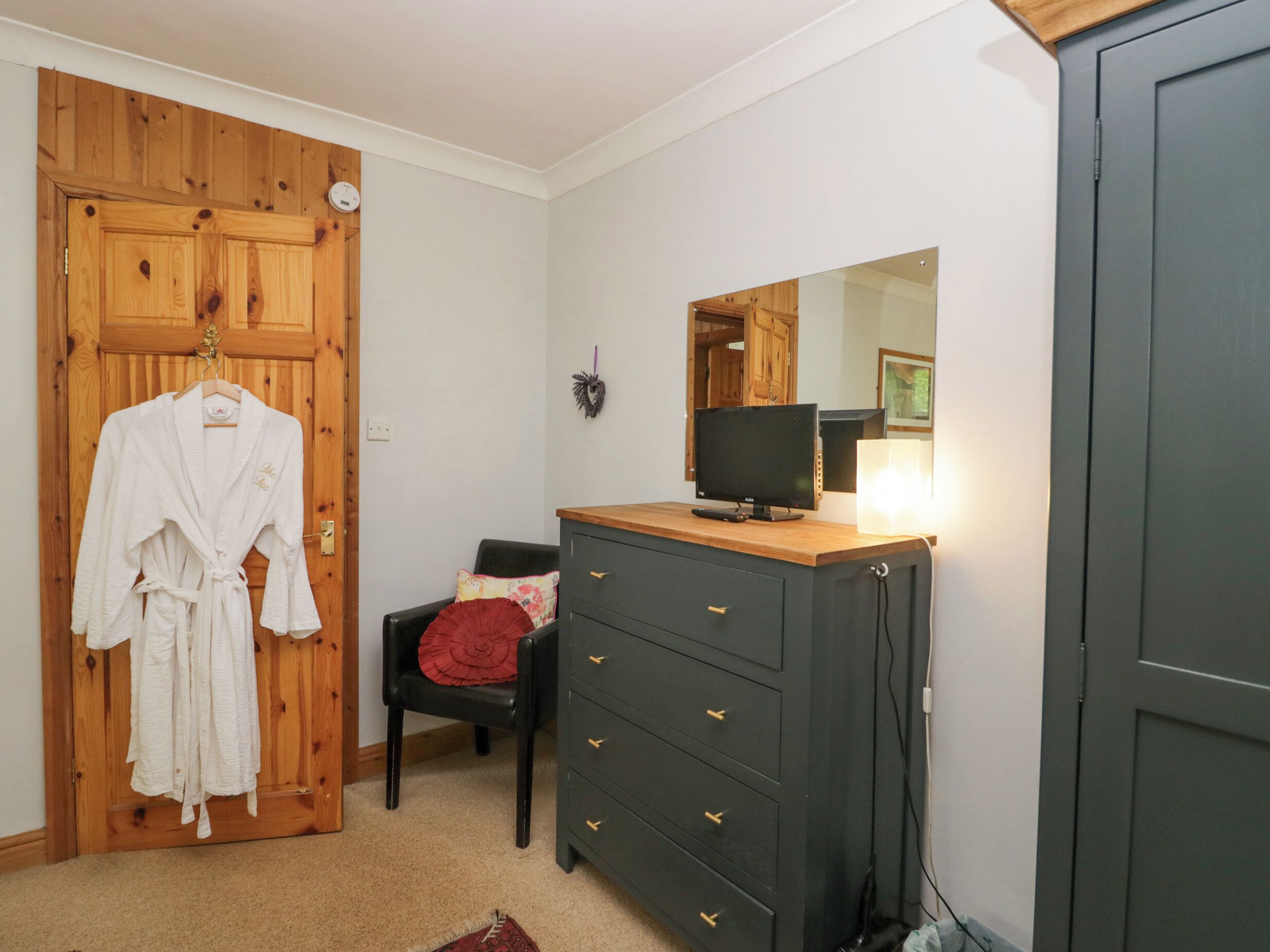 Ein Zimmer mit einer Holztür, einem schwarzen Stuhl mit rotem Kissen, einem Holzkleiderschrank mit Spiegel und einem weißen Bademantel, der an der Tür hängt.