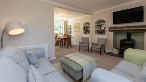 Un salon comprenant un canapé, un pouf et une cheminée. Un téléviseur est fixé au mur, et il y a également un coin repas avec des chaises et une table.