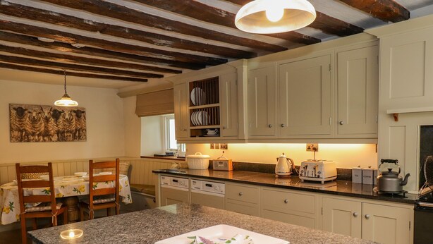 A kitchen with granite countertops, white cabinets, and exposed wooden beams. There is a dining table with chairs and a painting on the wall. - Low Cartmell Fold (Kendal)