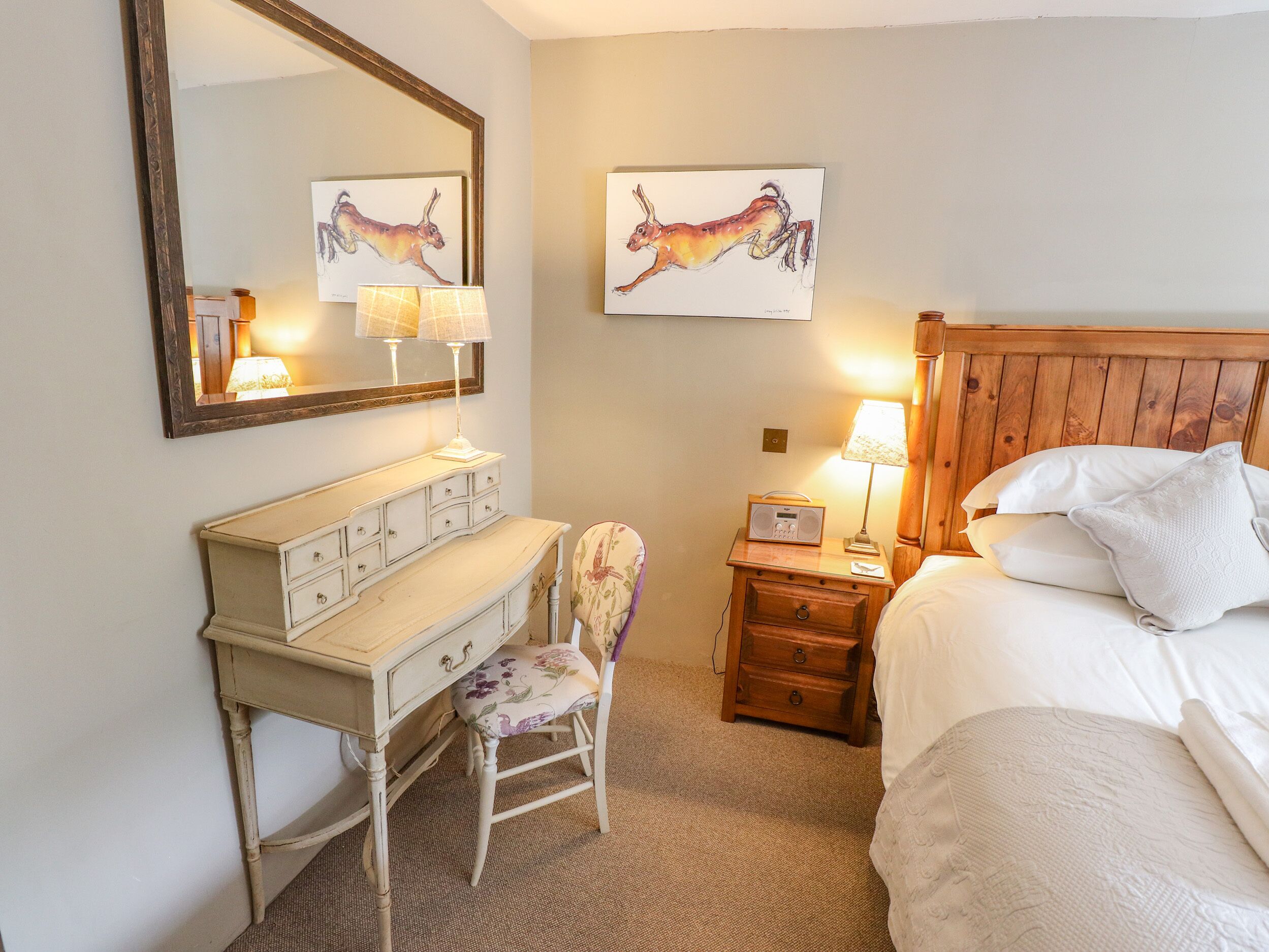 Habitación de hotel con una cama de madera, un tocador blanco, una silla, una mesita de noche con lámpara y dos cuadros colgados en la pared.