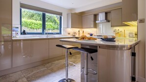 A modern kitchen with a large window, a central island with stools, and a view of greenery outside. - The Old Police House (Ambleside)