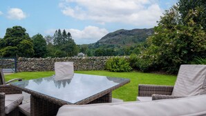 A patio with wicker furniture and a glass table, surrounded by a stone wall and lush greenery, with a mountain in the background. - The Old Police House (Ambleside)