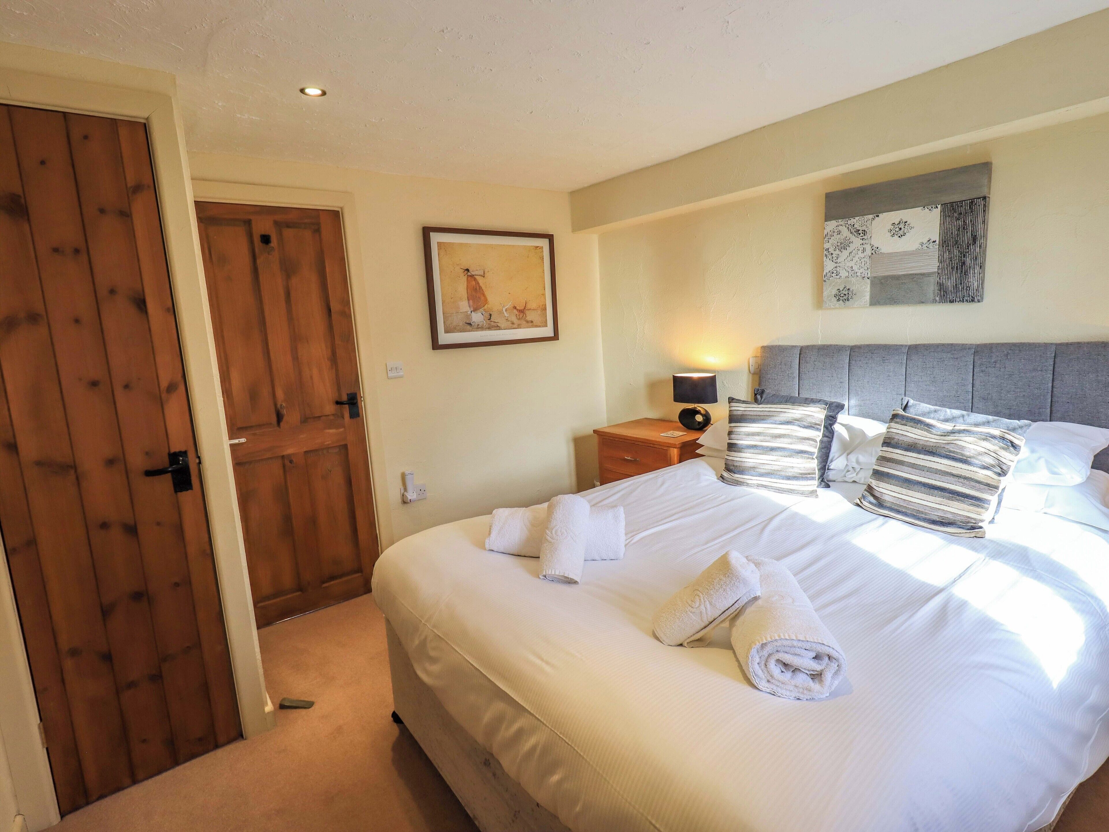 A neatly made bed with white linens and rolled towels, a bedside table with a lamp, a framed picture on the wall, and a wooden door.