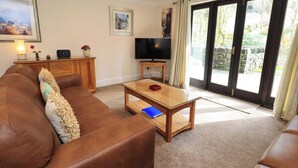 A living room with a brown leather sofa, a glass coffee table, a TV on a stand, and a view of the outdoors through large sliding glass doors. - Gillybeck (Ambleside)