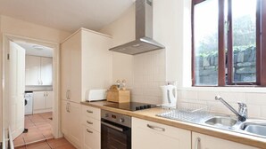 Une cuisine avec des armoires blanches, une hotte de cuisinière en acier inoxydable, un four encastré, un évier avec un égouttoir et une fenêtre donnant sur un espace verdoyant.