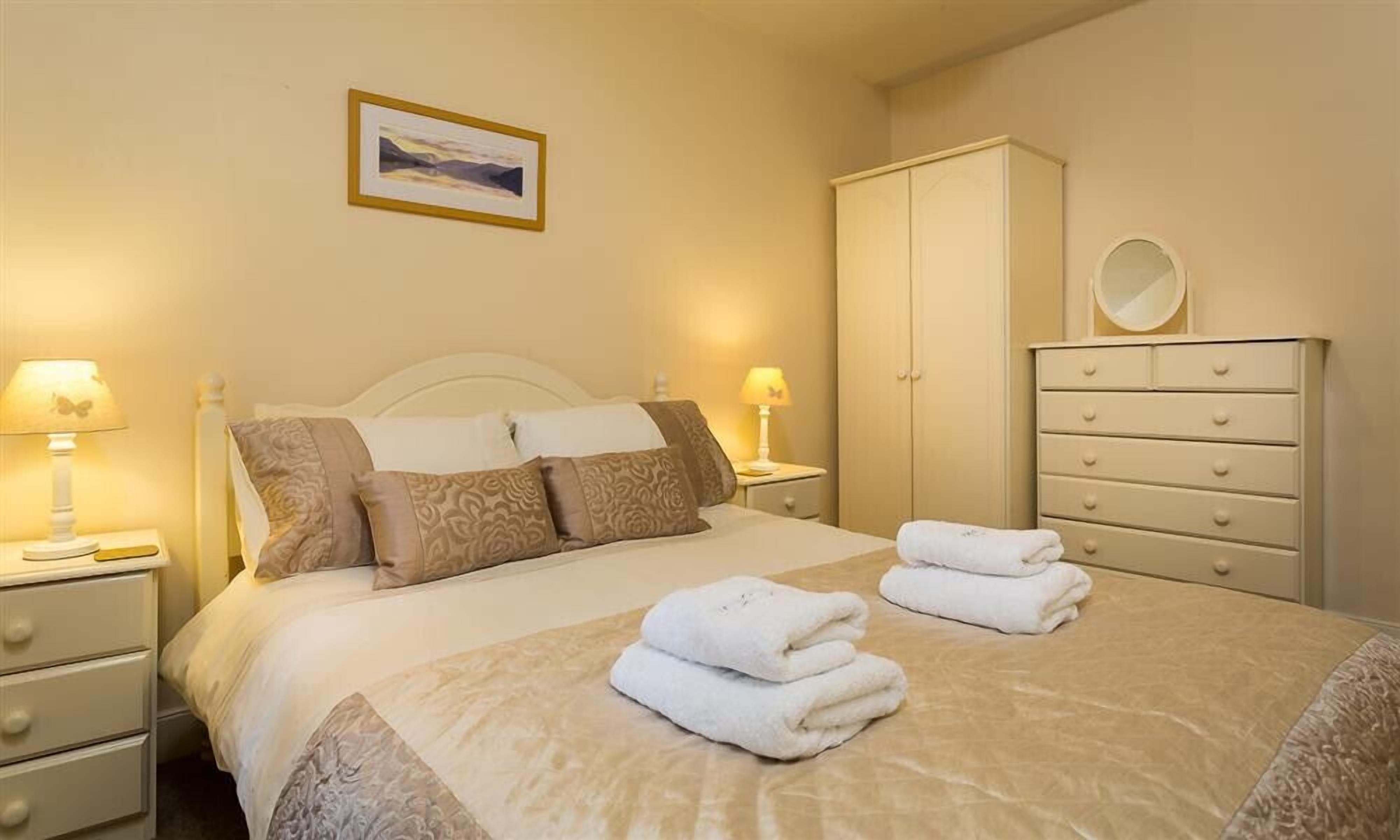 A neatly made bed with pillows, a bedside table with a lamp, a dresser with drawers, and a framed picture on the wall.