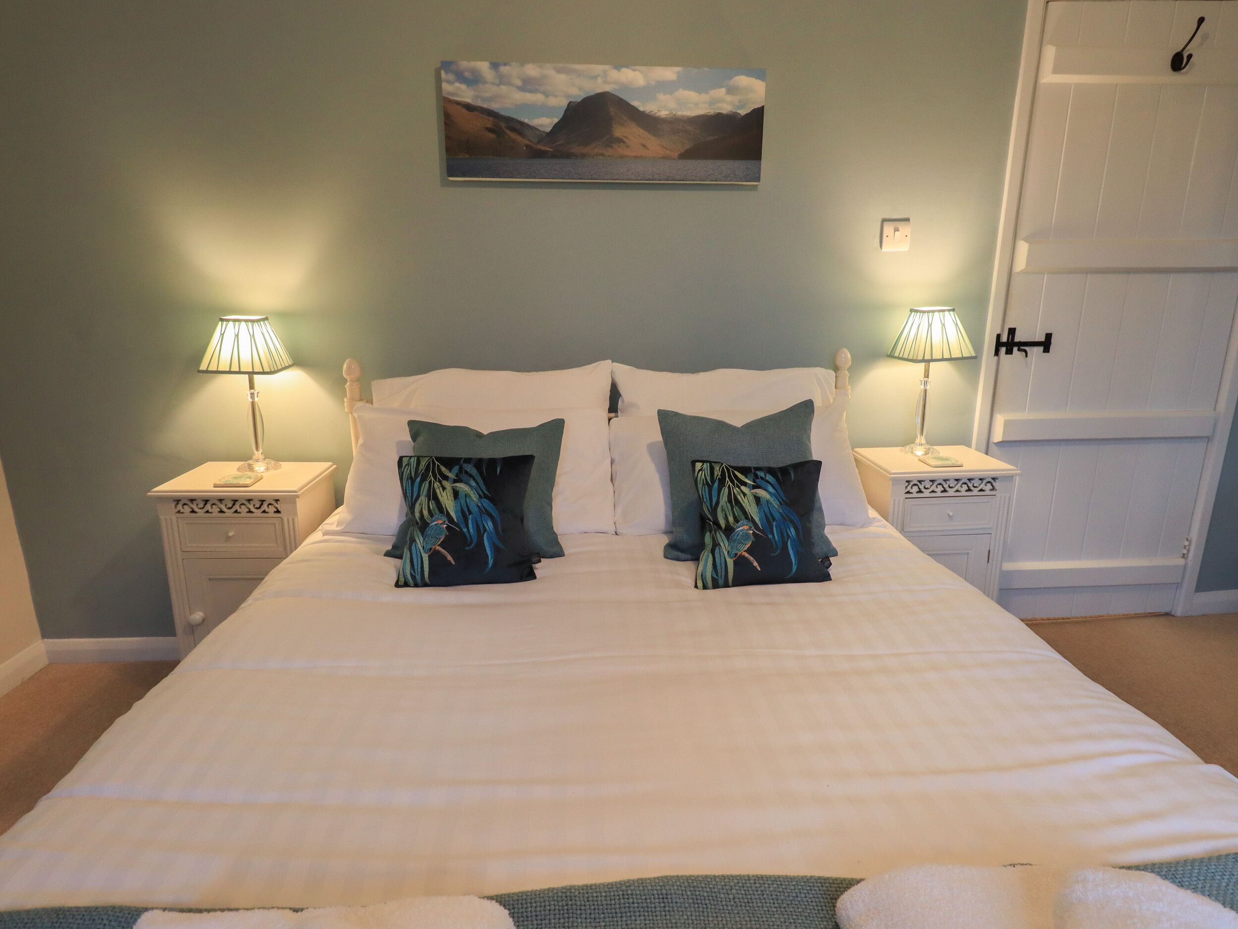 A neatly made bed with white linens and two decorative pillows, flanked by two bedside tables with lamps, in a room with a light blue wall and a painting above the bed.