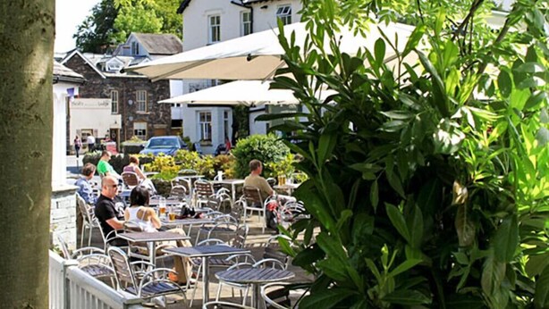 En udendørs café med borde og stole, omgivet af grøn vegetation og bygninger.