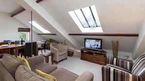 En loftstue med skrå vægge, ovenlysvinduer, et fjernsyn, en sofa, en lænestol og et spiseområde med bord og stole.