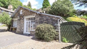 A stone house with a white door and windows, surrounded by greenery and a paved driveway.