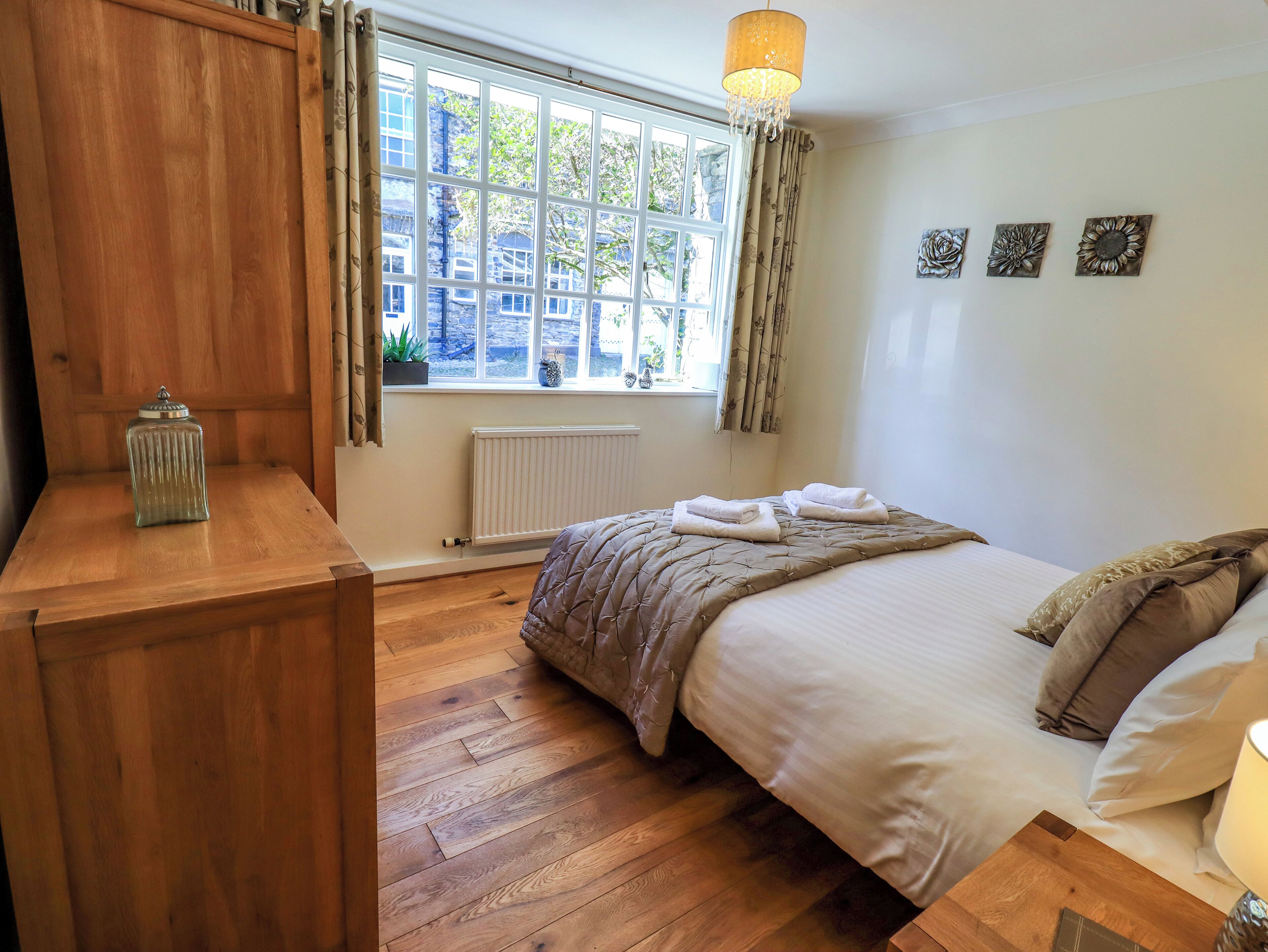 Une chambre avec une commode en bois, un lit recouvert d’une courtepointe beige, une fenêtre avec des rideaux et un radiateur.
