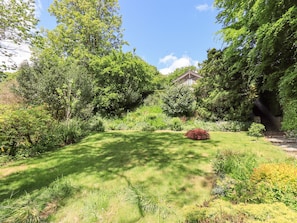 Um jardim exuberante com diversas plantas e árvores, um gramado bem cuidado e um pequeno edifício ao fundo.