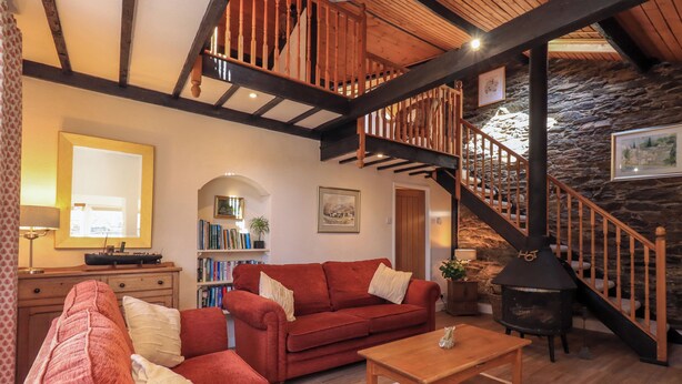 A cozy living room with a red sofa, a wooden coffee table, and a stone wall. A wooden staircase with a black railing leads to an upper level.
