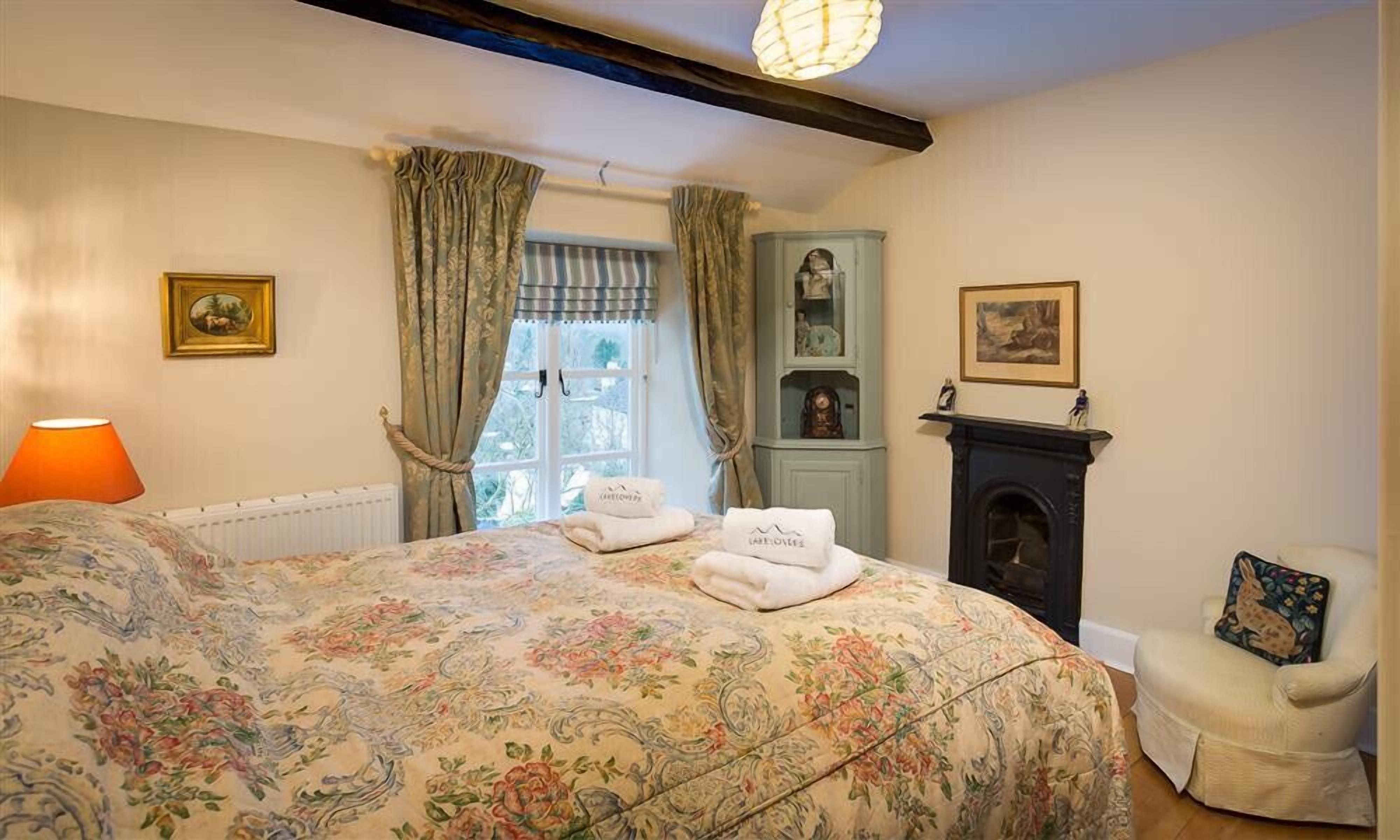 A bedroom with a floral bedspread, a fireplace, a chair with a cushion, and a window with curtains.