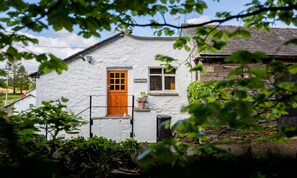 A white building with a wooden door and a small window, surrounded by greenery. - Barn End (Ambleside)