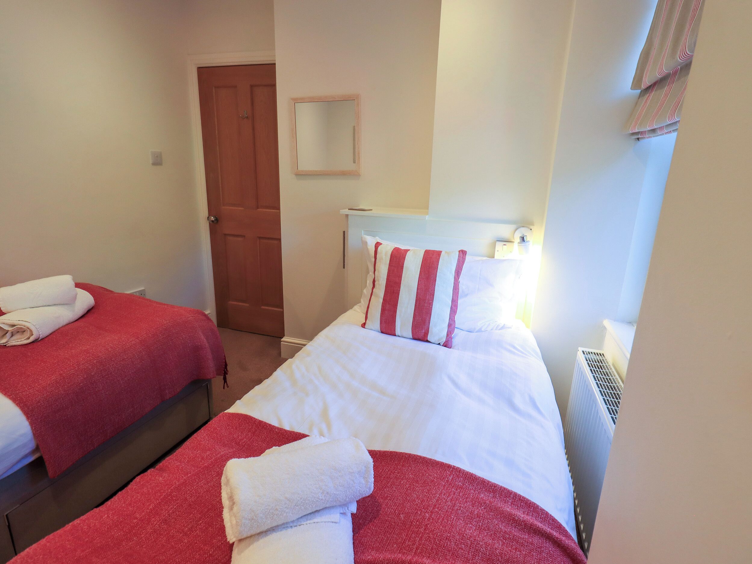 Habitación de hotel con dos camas, cada una con ropa de cama roja y blanca, una puerta de madera, un espejo pequeño y un radiador.