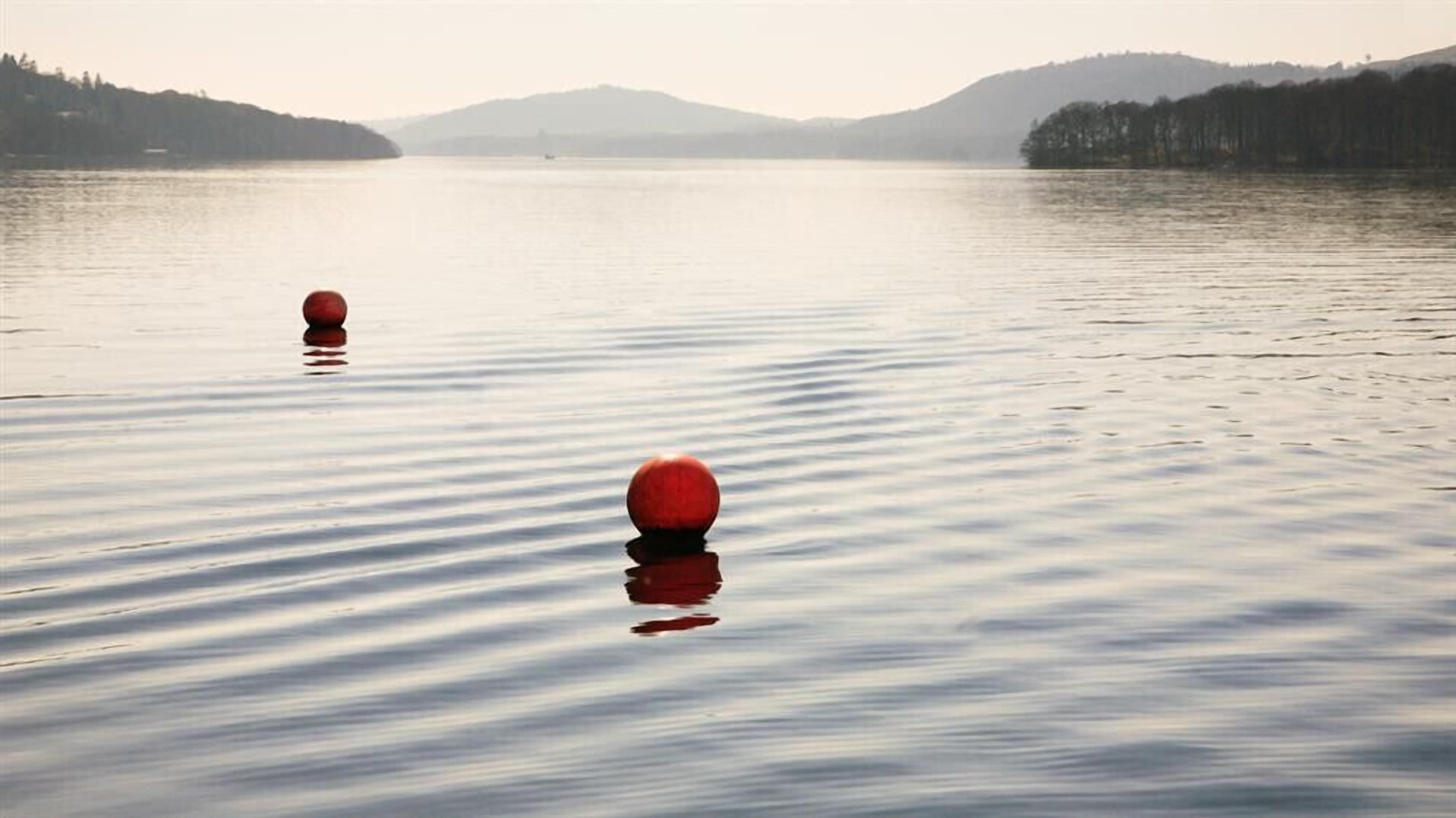 A calm lake with two red buoys in the foreground and a forested hill in the background.