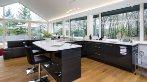 A modern kitchen with a central island, dark cabinetry, and large windows offering views of trees. - Tall Trees (Windermere)