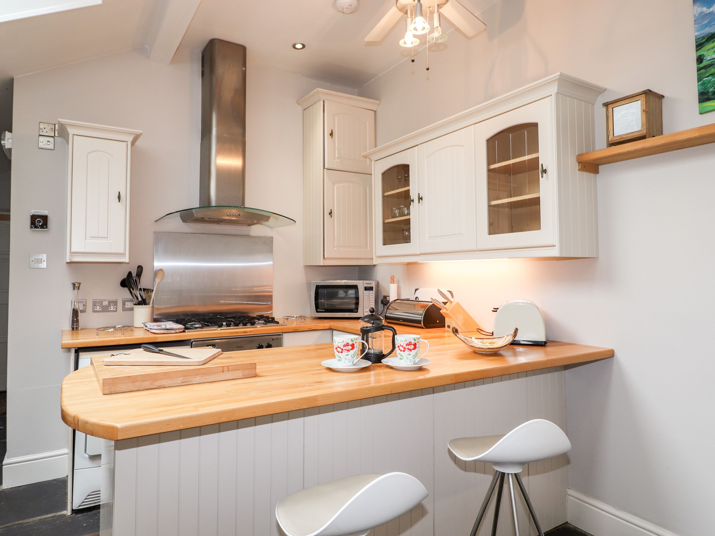 A modern kitchen with white cabinetry, a stainless steel range hood, a wooden countertop, and two white bar stools.