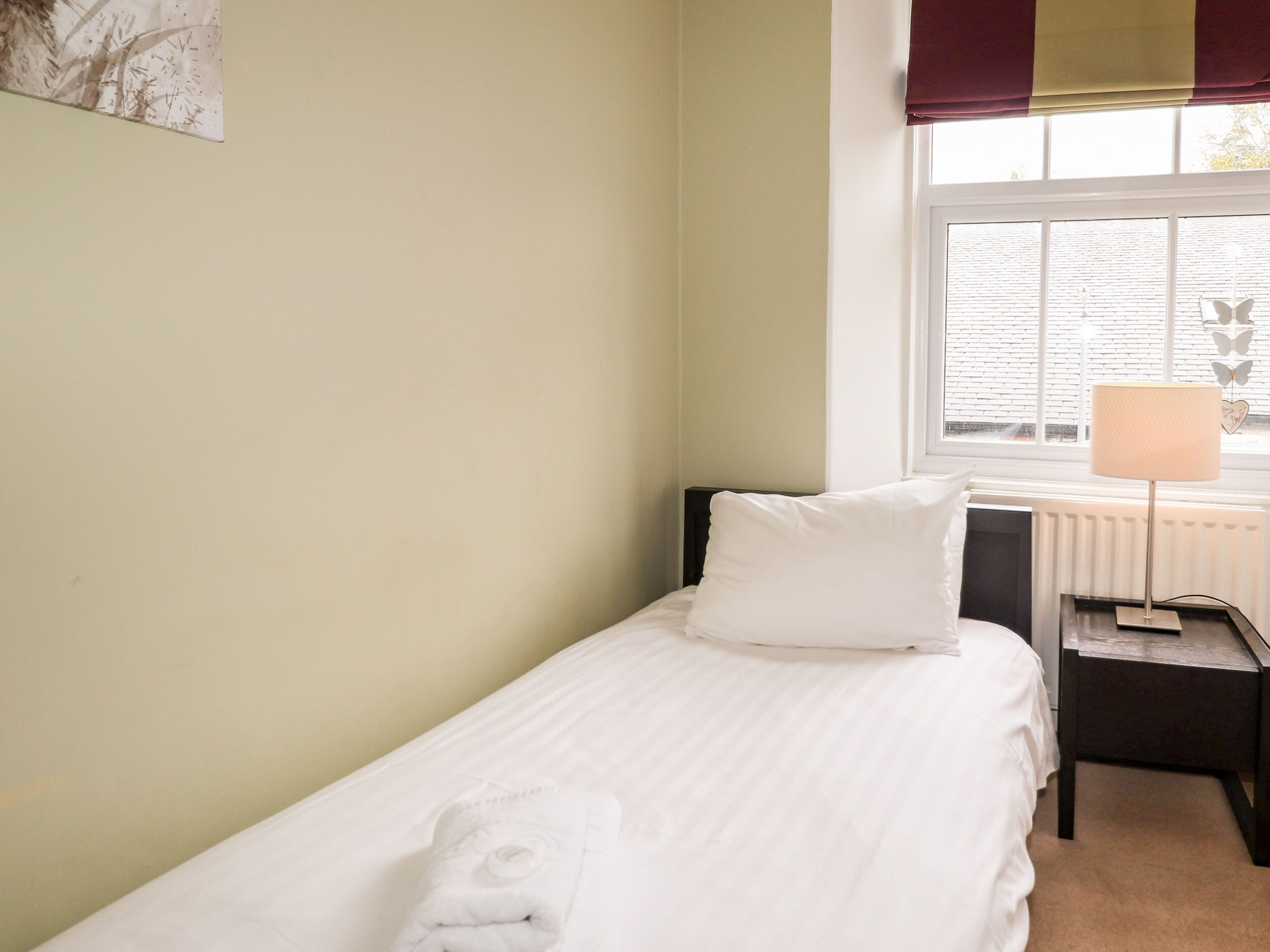 A single bed with a white pillow and a towel, a bedside table with a lamp, and a window with blinds.