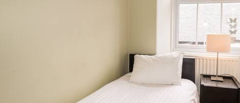 A single bed with a white pillow and a towel, a bedside table with a lamp, and a window with blinds.