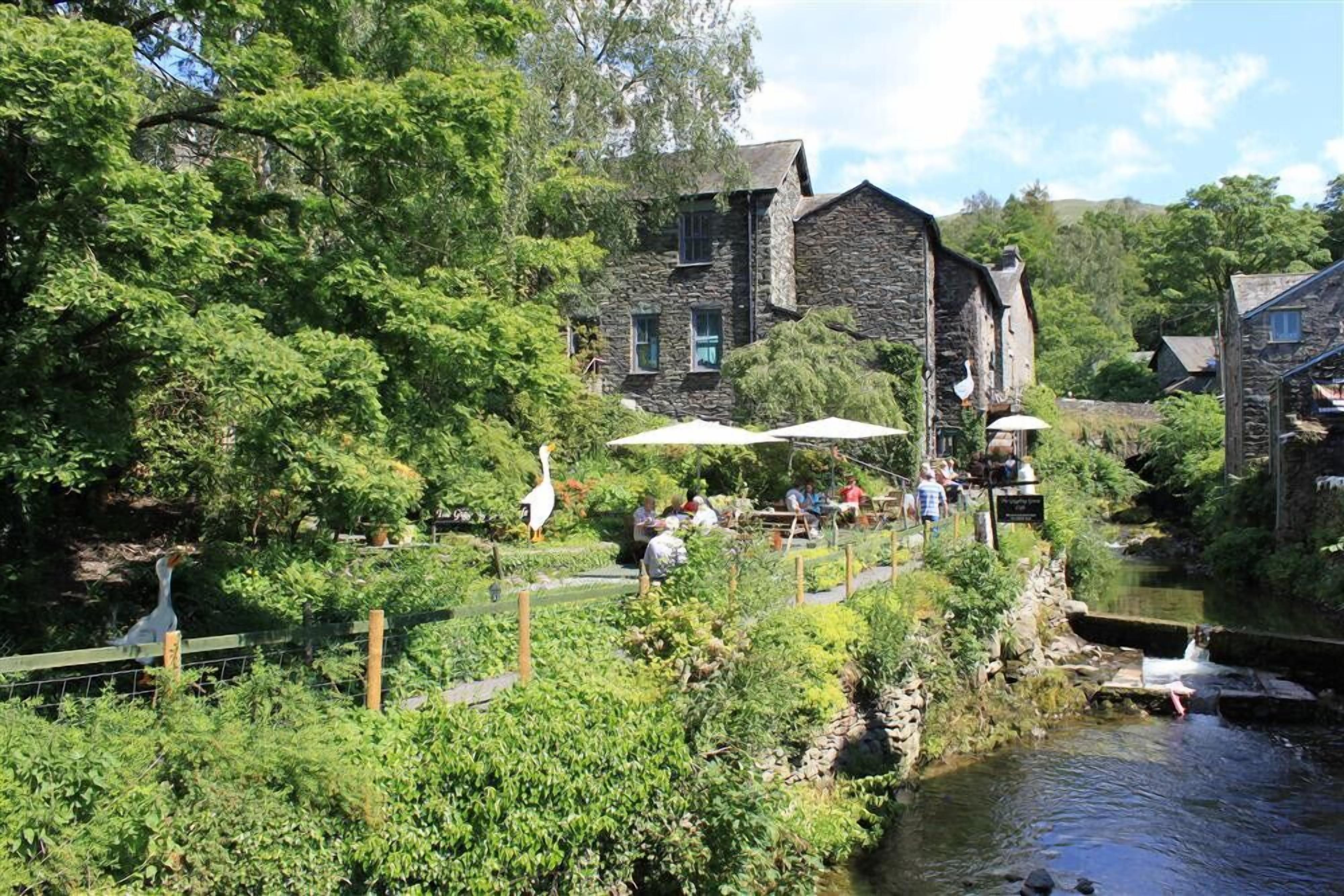 A riverside building with stone walls, surrounded by lush greenery and a small stream.