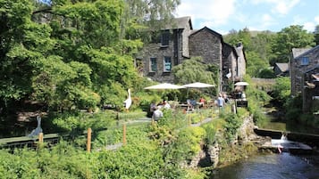 A riverside building with stone walls, surrounded by lush greenery and a small stream.