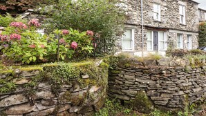 Une maison en pierre avec une porte blanche, entourée d’une végétation luxuriante et de fleurs roses.