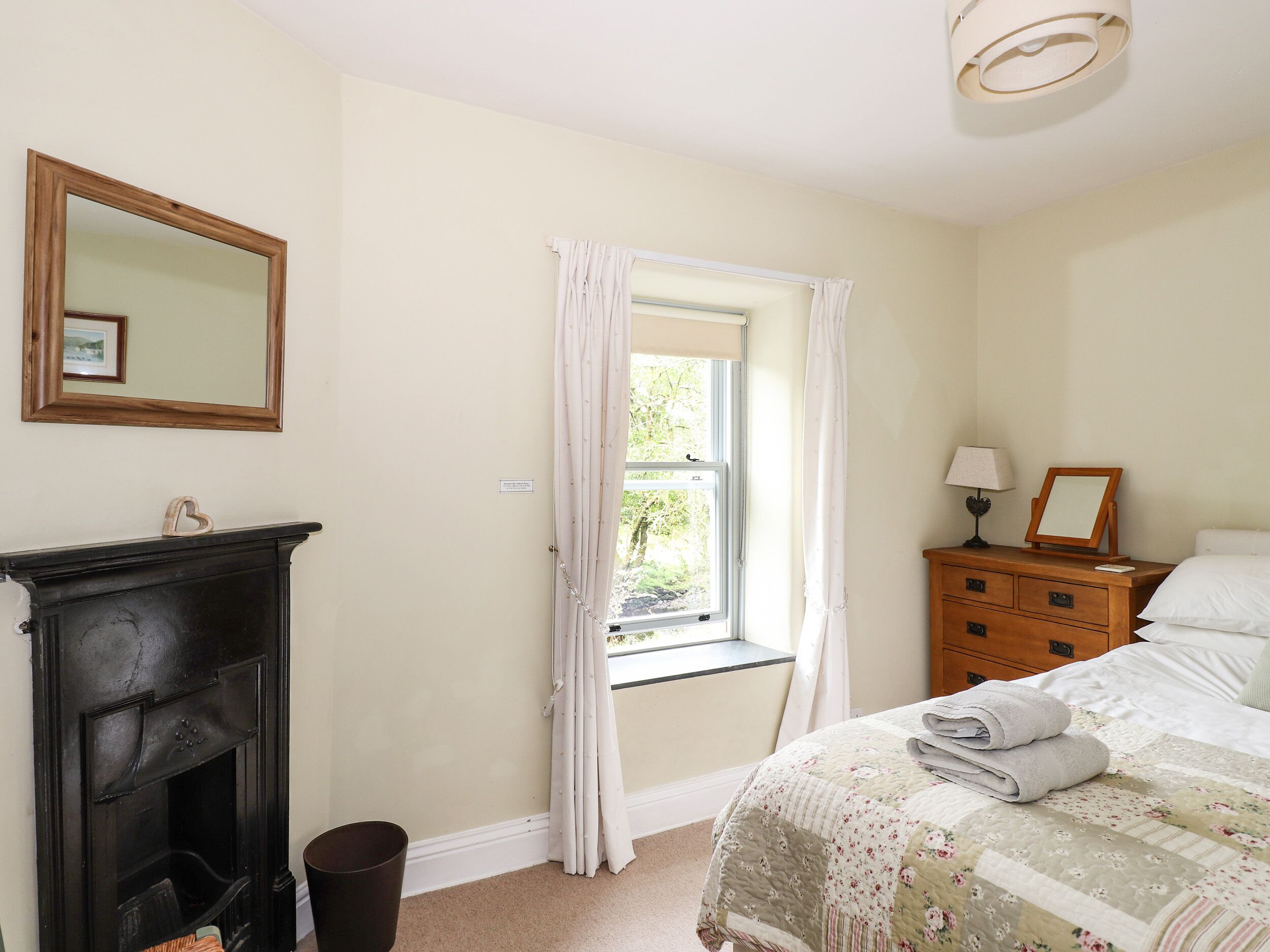 A bedroom with a bed, a dresser, a mirror, a fireplace, and a window with curtains.