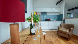 A modern living room with a wooden floor, a red lamp, a table with a bottle and glasses, a chair, and a kitchen in the background. - Old Sawrey Stores (Ambleside)