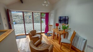 A cozy living room with a stone wall, a TV, a dining table with chairs, and a fireplace. - Old Sawrey Stores (Ambleside)