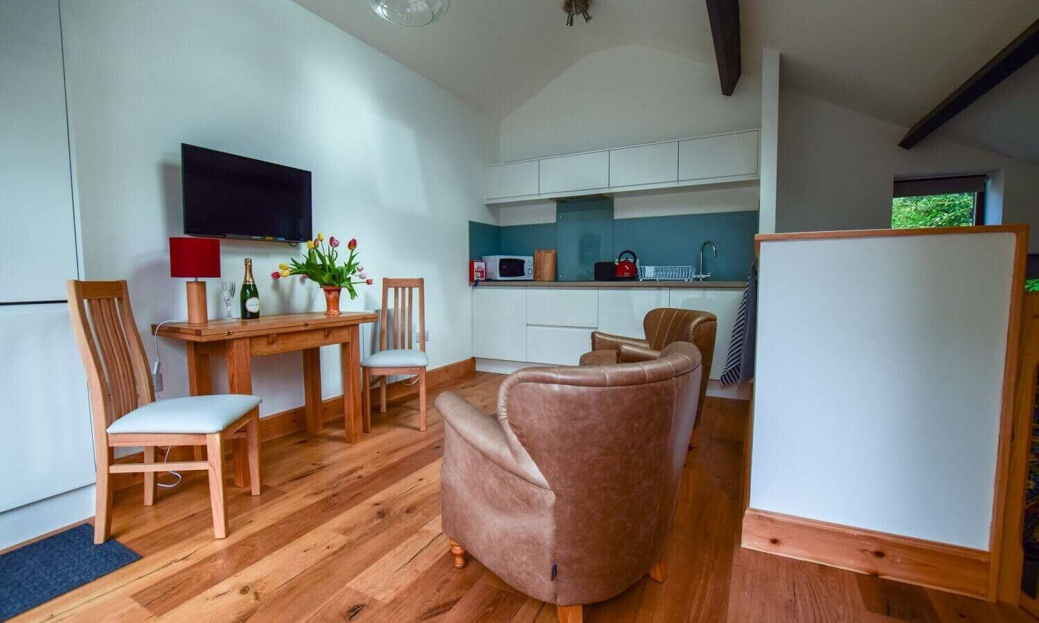 A compact living space with a wooden dining table, two chairs, a sofa, a television, a kitchen area with cabinets, and a refrigerator.