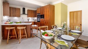 A modern kitchen with wooden cabinets, a central island, and a dining area with a glass table set for four. - Pippin Cottage (Keswick)
