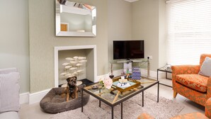 A living room with a dog bed, a glass coffee table with books and a wine glass, a television, and an orange patterned armchair. - Pippin Cottage (Keswick)