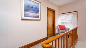 A hallway with a wooden door, a framed painting, a window with blinds, and a grey chair with a red cushion. - Pippin Cottage (Keswick)