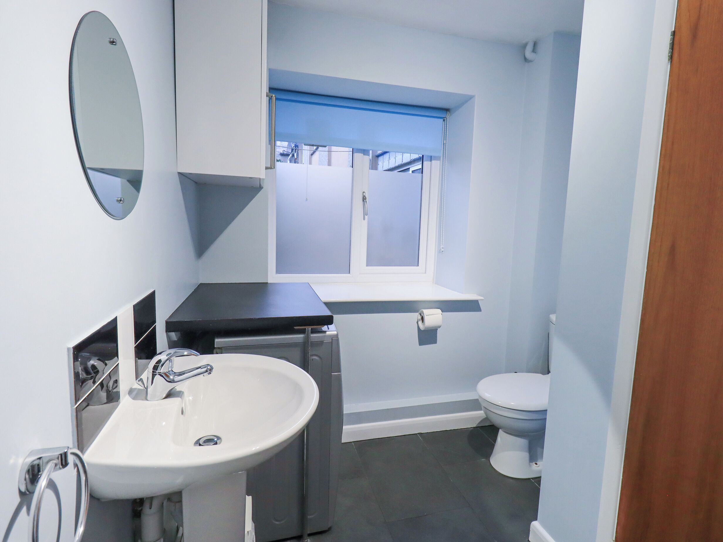 Ein kleines Badezimmer mit einem weißen Waschbecken, einem Spiegel, einer Toilette und einem Fenster mit blauer Jalousie.