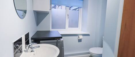 Ein kleines Badezimmer mit einem weißen Waschbecken, einem Spiegel, einer Toilette und einem Fenster mit blauer Jalousie.