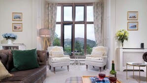 A cozy living room with a brown leather sofa, two white armchairs, a coffee table with a tray of wine glasses and a bottle, and a view of a scenic landscape through large windows. - Brunt How (Ambleside)