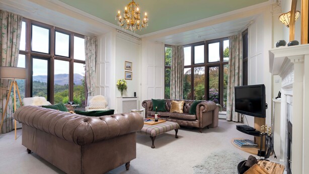 A spacious living room with a brown leather sofa, a matching armchair, and a green ottoman. There is a television on a stand, a fireplace, and a large window offering a view of the outdoors. - Brunt How (Ambleside)