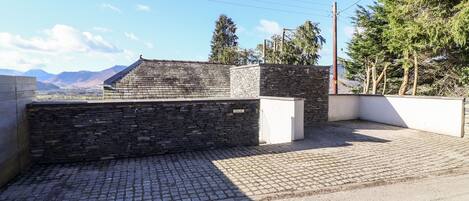 A stone building with a sloped roof, a paved driveway, and a view of mountains in the background.