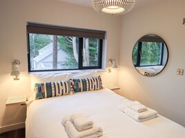 A neatly made bed with white linens and folded towels, a large window with a view of trees, and a round mirror on the wall.