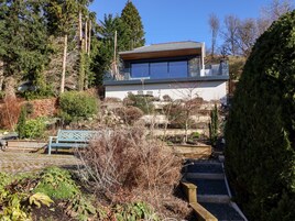 A modern house with a large window, surrounded by a garden with various plants and a wooden bench.