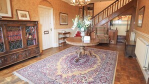 A spacious room with a wooden floor, a large ornate rug, a round table with a vase of flowers, a wooden cabinet with glass doors, a plaid armchair, and a staircase leading to an upper level.