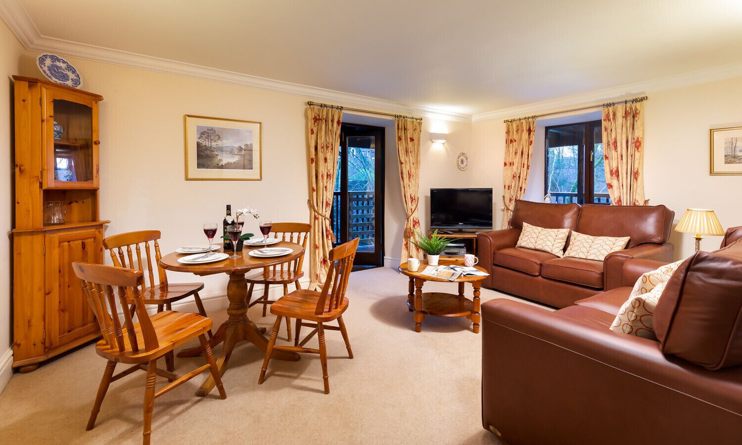 A living room with a brown leather sofa, wooden dining table with chairs, a television, and a cabinet.