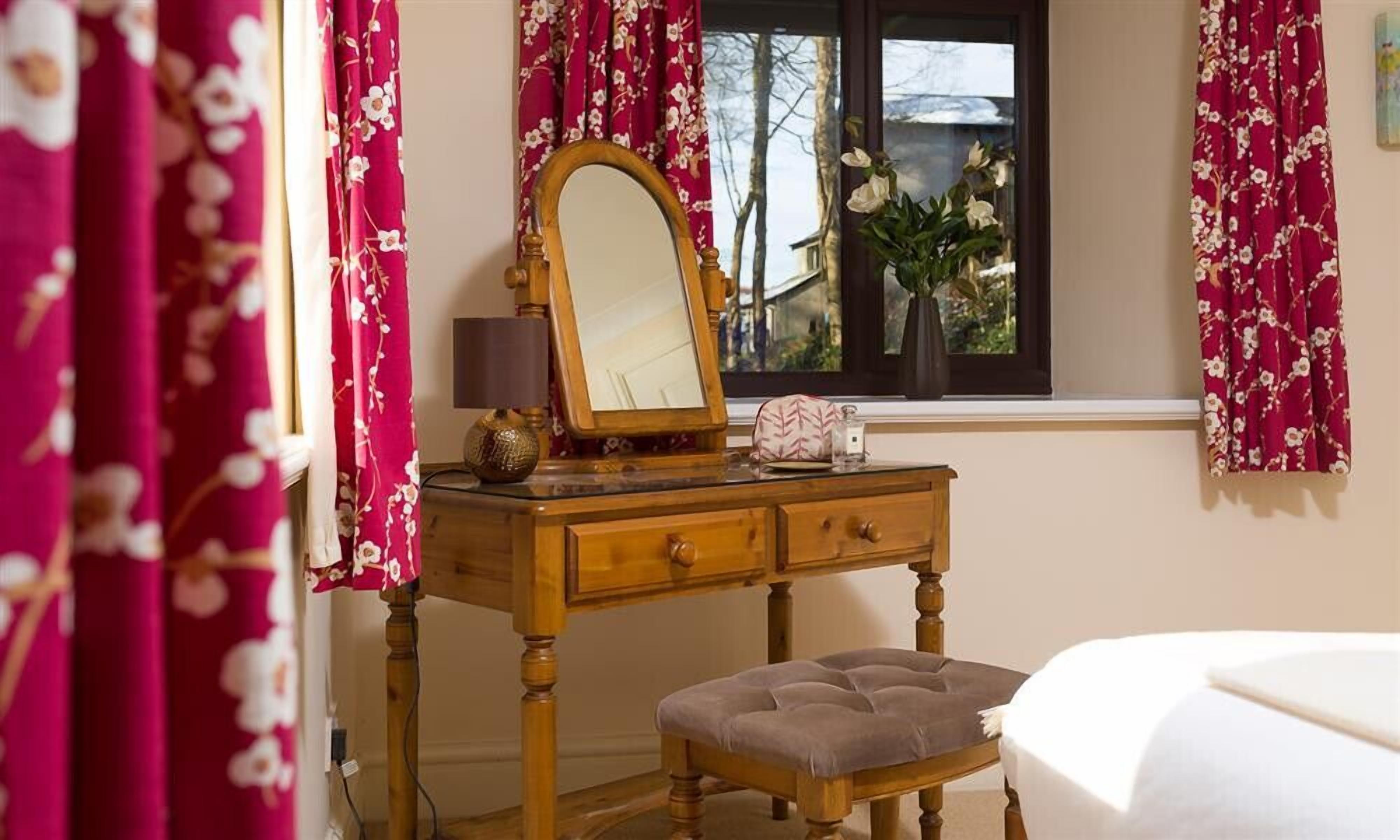 Une coiffeuse en bois, avec un miroir, une lampe et une petite table. Il y a une fenêtre donnant sur des arbres, et un vase rempli de fleurs est posé sur le rebord de la fenêtre. Un banc est placé devant la coiffeuse.